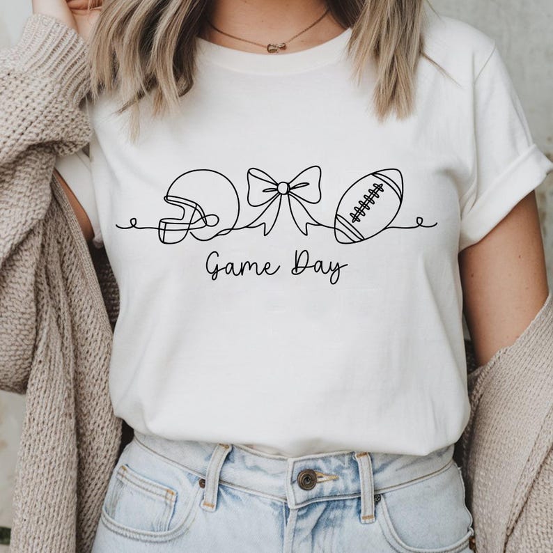 Pode incluir: Camiseta creme com um desenho a tra&ccedil;o preto de um capacete de futebol americano, um la&ccedil;o e uma bola de futebol. As palavras "Game Day" est&atilde;o escritas abaixo. A camisa &eacute; usada com jeans azuis.