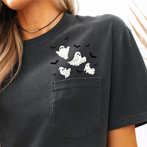 May include: Dark gray t-shirt featuring a pocket and a graphic of white ghosts and black bats. The shirt has a classic crew neck and short sleeves, perfect for Halloween or spooky season.