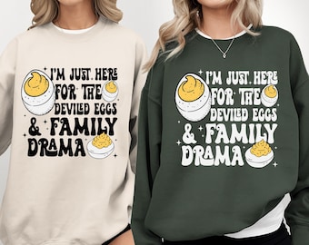 May include: Two sweatshirts, one cream and one dark green, with the text "I'M JUST HERE FOR THE DEVILED EGGS & FAMILY DRAMA". The design includes illustrations of deviled eggs in a retro style.