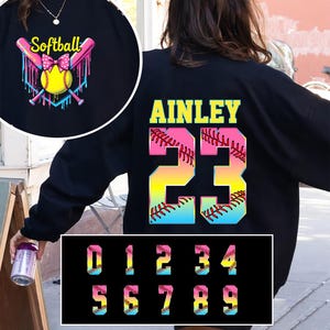 Pu&ograve; includere: Felpa nera con un design a forma di palla da softball e il nome "AINLEY" e il numero "23" sul retro. I numeri sono arcobaleno con un design a punto di softball. La parte anteriore della felpa ha una palla da softball con un fiocco rosa e vernice che cola. I numeri da 0 a 9 sono mostrati in colori arcobaleno con un design a punto di softball.
