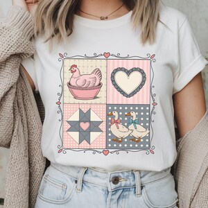 以下が含まれることがあります： パッチワークデザインの白いTシャツ。ボウルに入った鶏、ハート、キルトブロック、リボンをつけた2羽のアヒルがデザインされています。デザインは装飾的なボーダーで縁取られ、ピンク、グレー、白の色を使用しています。