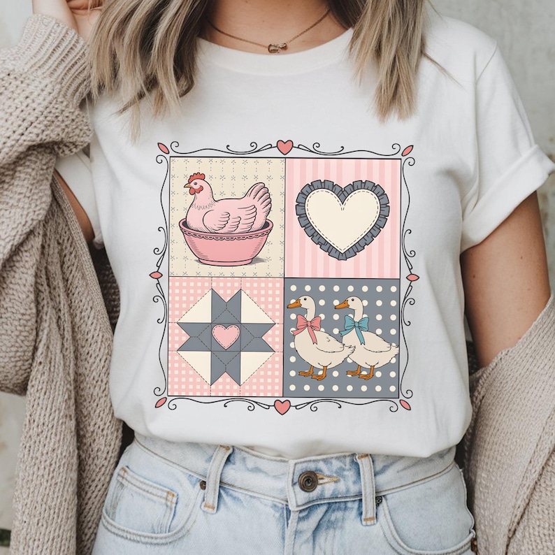 以下が含まれることがあります： パッチワークデザインの白いTシャツ。ボウルに入った鶏、ハート、キルトブロック、リボンをつけた2羽のアヒルがデザインされています。デザインは装飾的なボーダーで縁取られ、ピンク、グレー、白の色を使用しています。