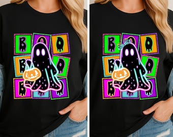 Puede incluir: Camiseta negra con un diseño de Halloween. El gráfico incluye un fantasma negro sosteniendo una calabaza, con la palabra "BOO" encima. Las letras y el fantasma están delineados en verde neón, rosa y azul.