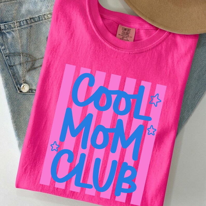 May include: A bright pink t-shirt with the words "Cool Mom Club" in blue script and block letters. The text is set against a background of pink vertical stripes. The shirt is folded on top of a pair of blue jeans.