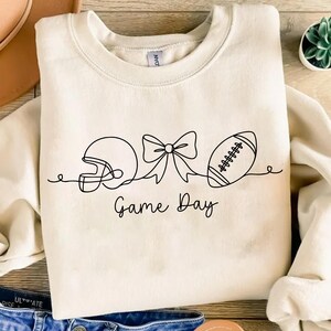 Pode incluir: Camisola creme com um desenho a tra&ccedil;o preto de um capacete de futebol americano, um la&ccedil;o e uma bola de futebol. As palavras "Game Day" est&atilde;o escritas em fonte de script por baixo. A camisola est&aacute; dobrada numa superf&iacute;cie clara.