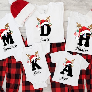 Könnte beinhalten: Weiße Familienpyjamas zum Weihnachtsthema mit rot-schwarz karierten Büffelhosen. Die Shirts zeigen ein Buchstabendesign mit Weihnachtsmütze und Rentiergeweih. Namen sind unter den Buchstaben aufgedruckt.