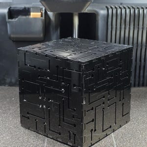 May include: A black cube with a glossy finish featuring intricate, recessed circuit board-like patterns on its exterior surfaces. The cube is positioned directly beneath the print head of a 3D printer, which is visible in the upper portion of the frame.