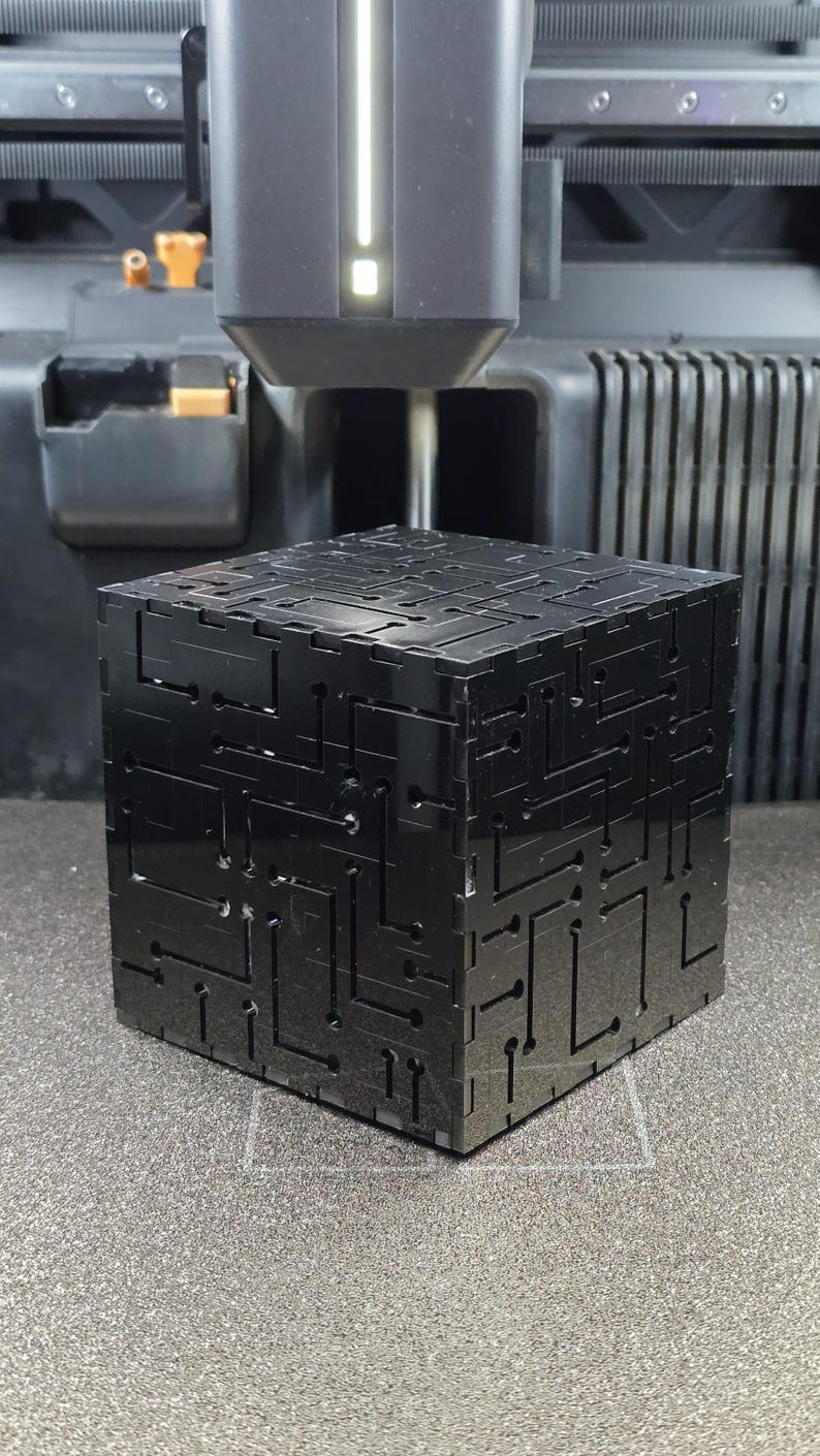 May include: A black cube with a glossy finish featuring intricate, recessed circuit board-like patterns on its exterior surfaces. The cube is positioned directly beneath the print head of a 3D printer, which is visible in the upper portion of the frame.