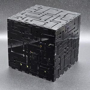 May include: A black cubic box constructed from interlocking panels featuring a laser-cut circuit board pattern on all visible exterior surfaces. The glossy material reveals small glimpses of internal components through the cutouts.