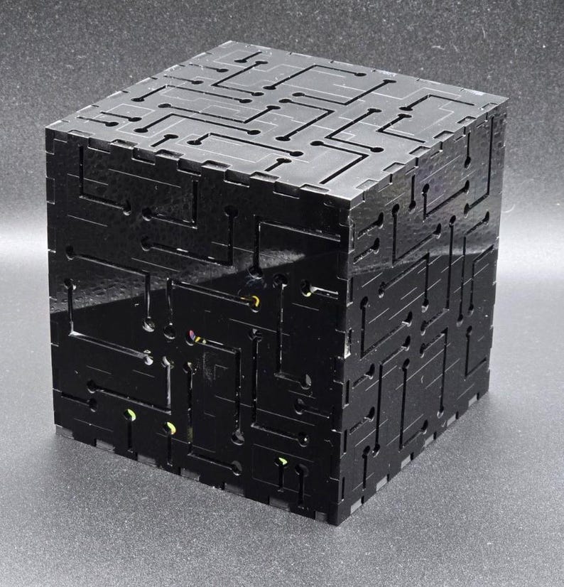 May include: A black cubic box constructed from interlocking panels featuring a laser-cut circuit board pattern on all visible exterior surfaces. The glossy material reveals small glimpses of internal components through the cutouts.