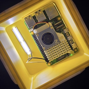 May include: A yellow plastic case with a green circuit board inside. The circuit board has a small fan and a heat sink. The text "Raspberry Pi not included" is printed below the case.