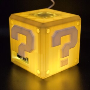 May include: A yellow cube-shaped lamp with a question mark on each side. The lamp is lit and glowing.