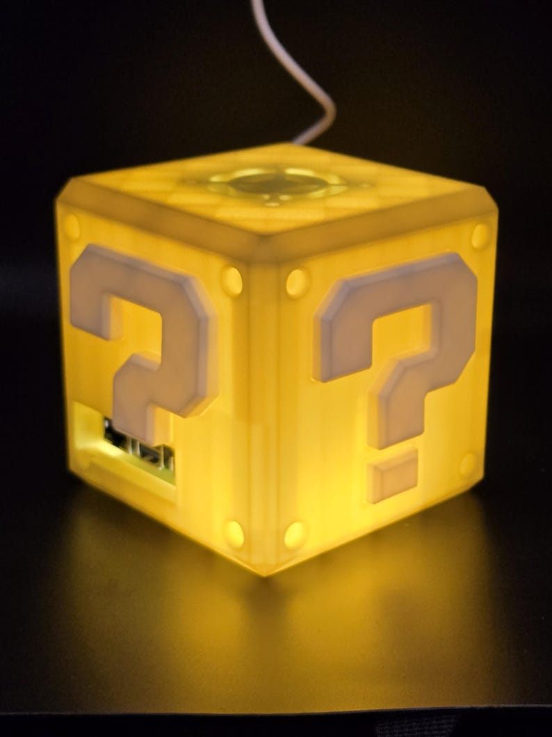 May include: A yellow cube-shaped lamp with a question mark on each side. The lamp is lit and glowing.