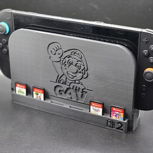 Puede incluir: Una consola Nintendo Switch negra se encuentra en un soporte personalizado. El soporte presenta un diseño de Mario y ranuras para cartuchos de juego. La palabra "GAY" es visible en el soporte.