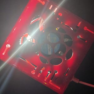 May include: A square red plastic housing with cut-out patterns and a central circular opening containing a brushless fan. The device is illuminated by a bright red light and features a white power cable connected to the side of the frame.