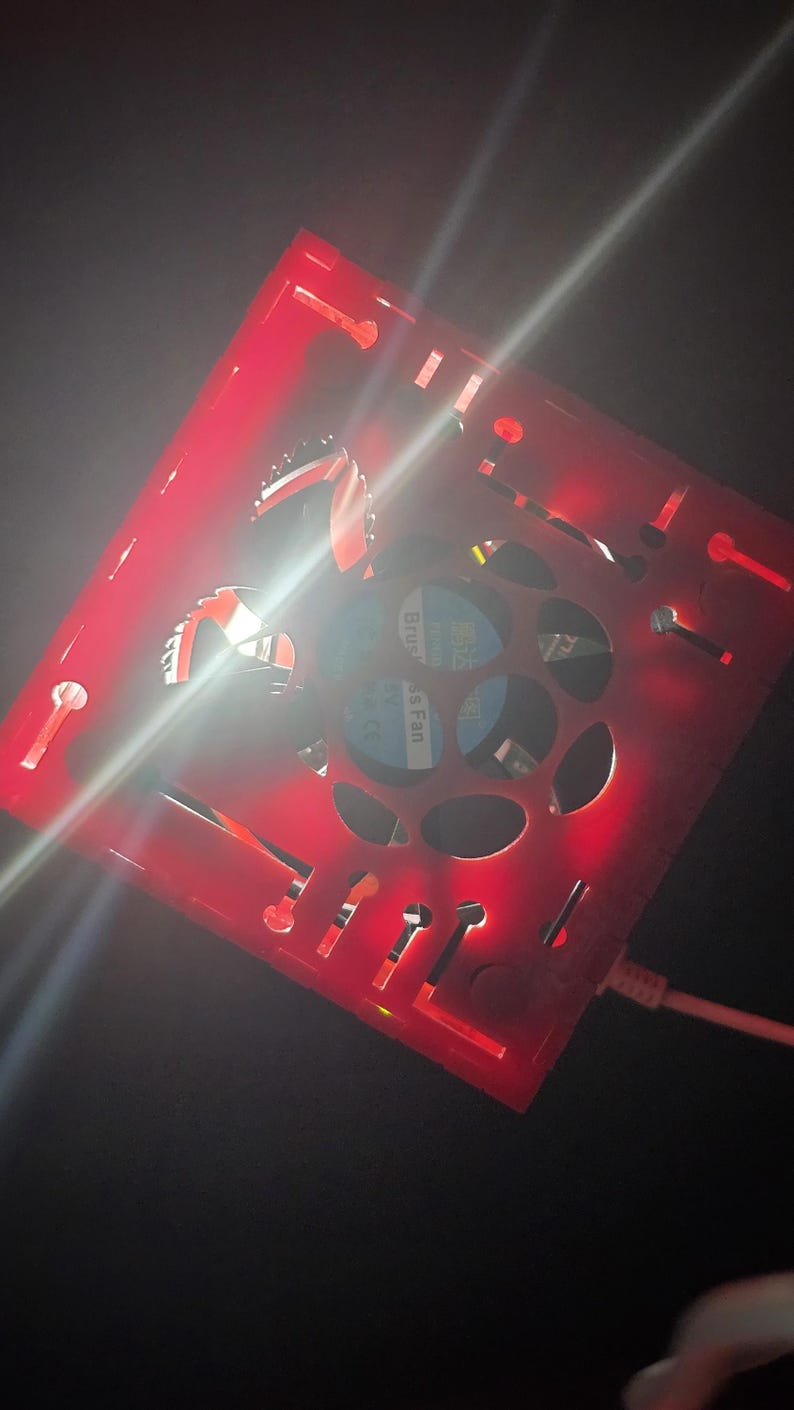 May include: A square red plastic housing with cut-out patterns and a central circular opening containing a brushless fan. The device is illuminated by a bright red light and features a white power cable connected to the side of the frame.