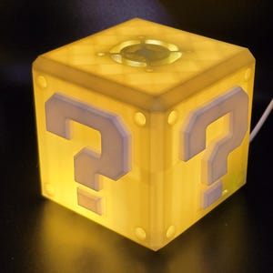 May include: A yellow cube-shaped night light with a question mark on each side. The light is on and glowing.