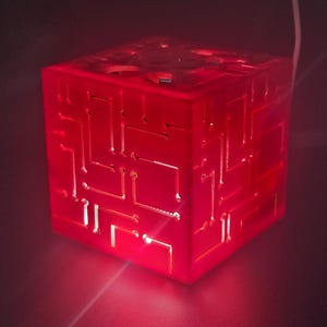 May include: A translucent red cube lamp featuring an intricate, recessed circuit board pattern on its exterior surfaces. The cube emits a vibrant red glow from within, highlighting the geometric lines and pathways etched into the material.