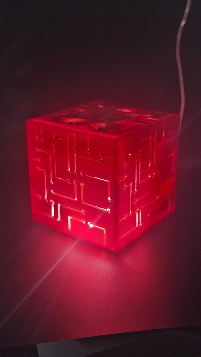 May include: A translucent red cube lamp featuring an intricate, recessed circuit board pattern on its exterior surfaces. The cube emits a vibrant red glow from within, highlighting the geometric lines and pathways etched into the material.