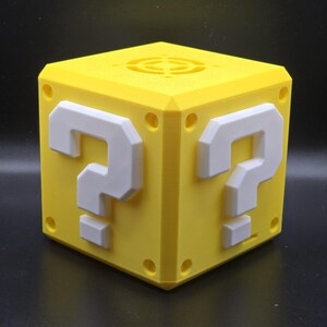 May include: A yellow 3D printed question mark block with white question marks on each side. The block has a circular vent on the top.