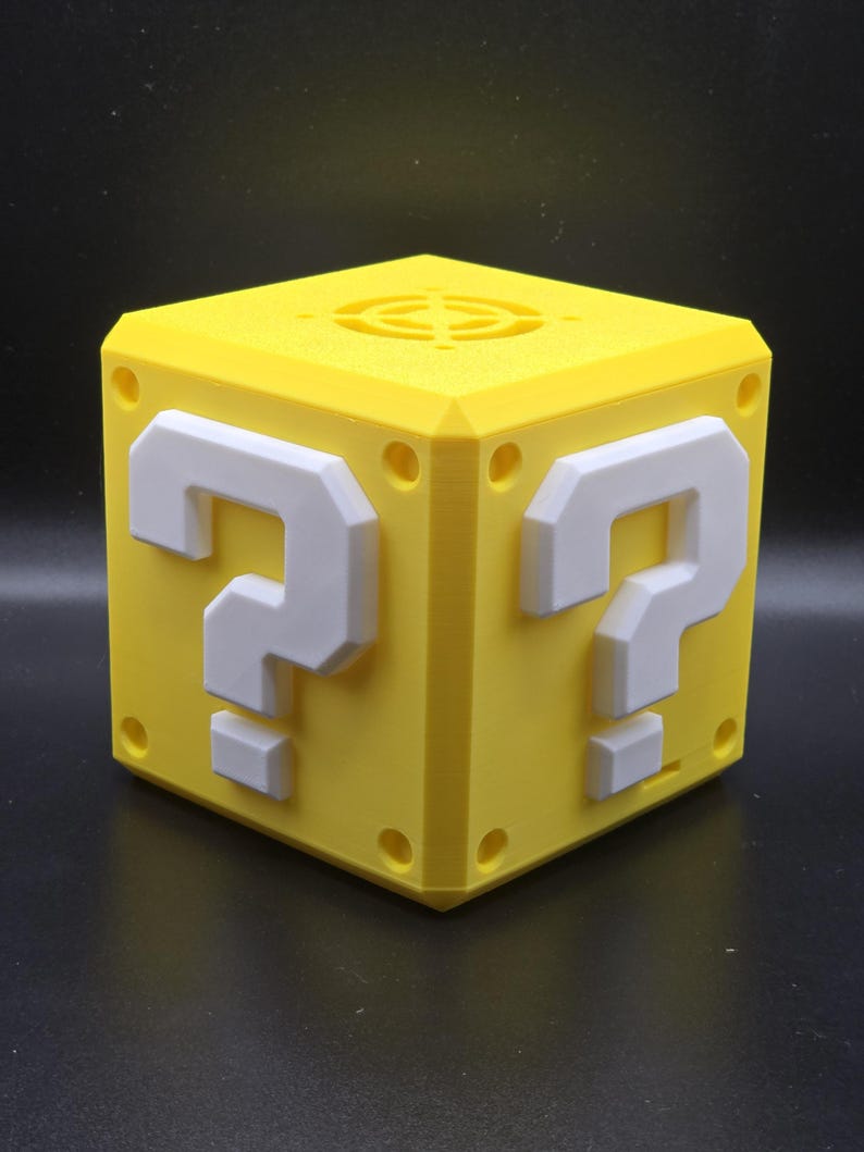 May include: A yellow 3D printed question mark block with white question marks on each side. The block has a circular vent on the top.