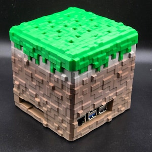 Könnte beinhalten: Ein 3D-gedrucktes Minecraft-Case für einen Raspberry Pi, mit einem grünen Grasblock oben und braunen Erdblöcken an den Seiten. Das Case hat mehrere Anschlüsse an der Seite, darunter USB und Ethernet.