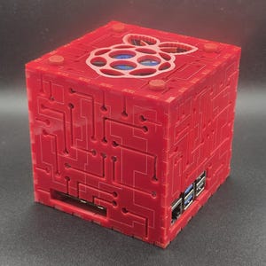 May include: A red, cube-shaped computer case featuring a laser-cut circuit board pattern on its exterior panels. The top panel displays a cutout of the Raspberry Pi logo, while the side panels reveal integrated USB and Ethernet ports.