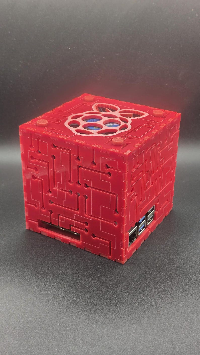 May include: A red, cube-shaped computer case featuring a laser-cut circuit board pattern on its exterior panels. The top panel displays a cutout of the Raspberry Pi logo, while the side panels reveal integrated USB and Ethernet ports.