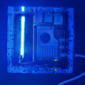 May include: A square, dark-colored enclosure containing a Raspberry Pi single-board computer with a cooling fan and heatsink. A bright blue LED light strip is mounted vertically inside the case, illuminating the internal components in blue.