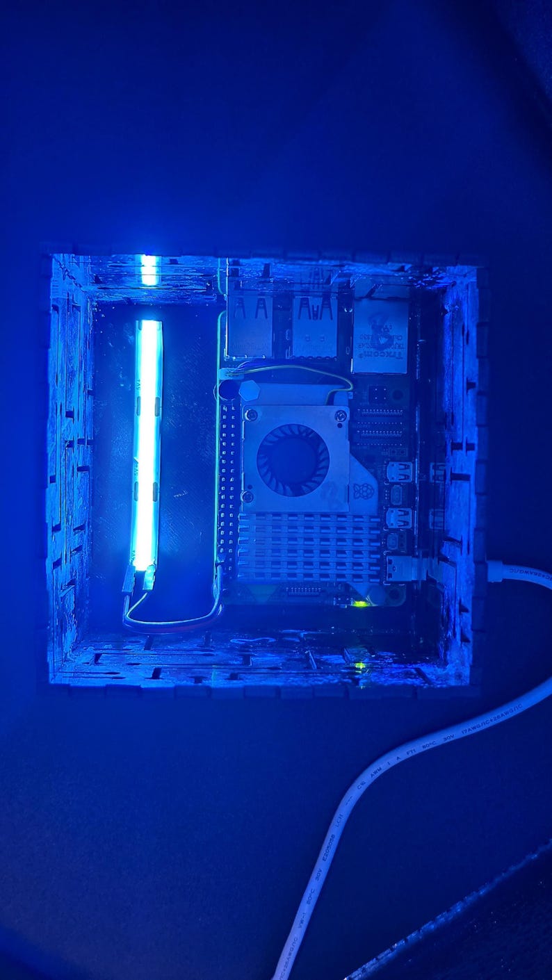May include: A square, dark-colored enclosure containing a Raspberry Pi single-board computer with a cooling fan and heatsink. A bright blue LED light strip is mounted vertically inside the case, illuminating the internal components in blue.