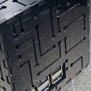 May include: A black, cubic enclosure featuring a laser-cut circuit board pattern on its exterior surfaces. The structure includes cutouts for an Ethernet port and multiple USB ports, revealing the internal electronic components within the housing.