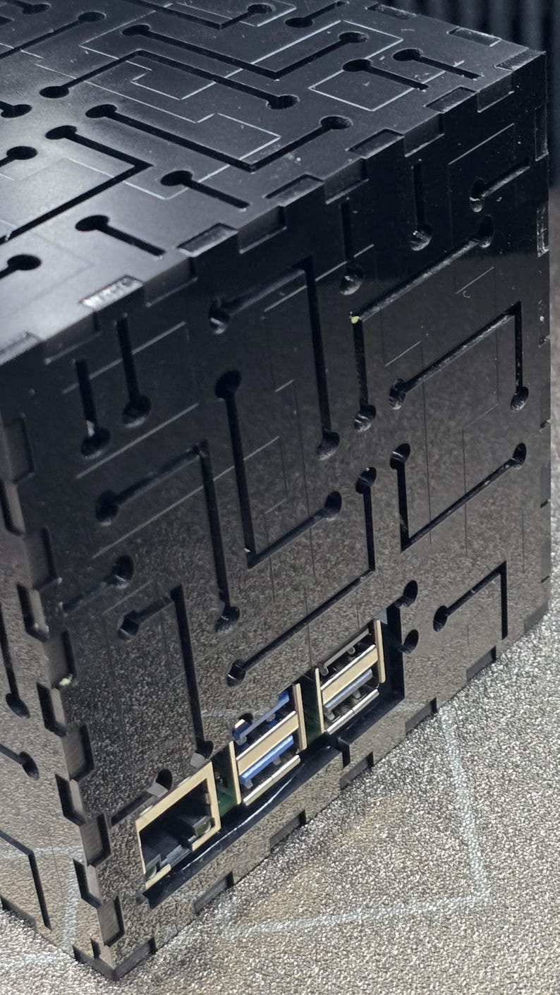 May include: A black, cubic enclosure featuring a laser-cut circuit board pattern on its exterior surfaces. The structure includes cutouts for an Ethernet port and multiple USB ports, revealing the internal electronic components within the housing.