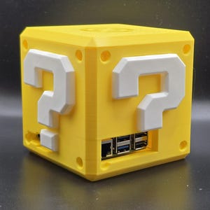 May include: A yellow 3D printed box with a question mark on each side. The box has a slot for a USB cable and a slot for a power cable.