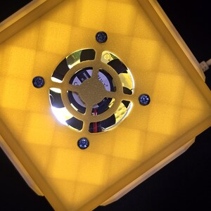 May include: A yellow square-shaped object with a white border and a black fan in the center. The fan has a white label with black text that reads "CUTL+ 24V 0.15A".