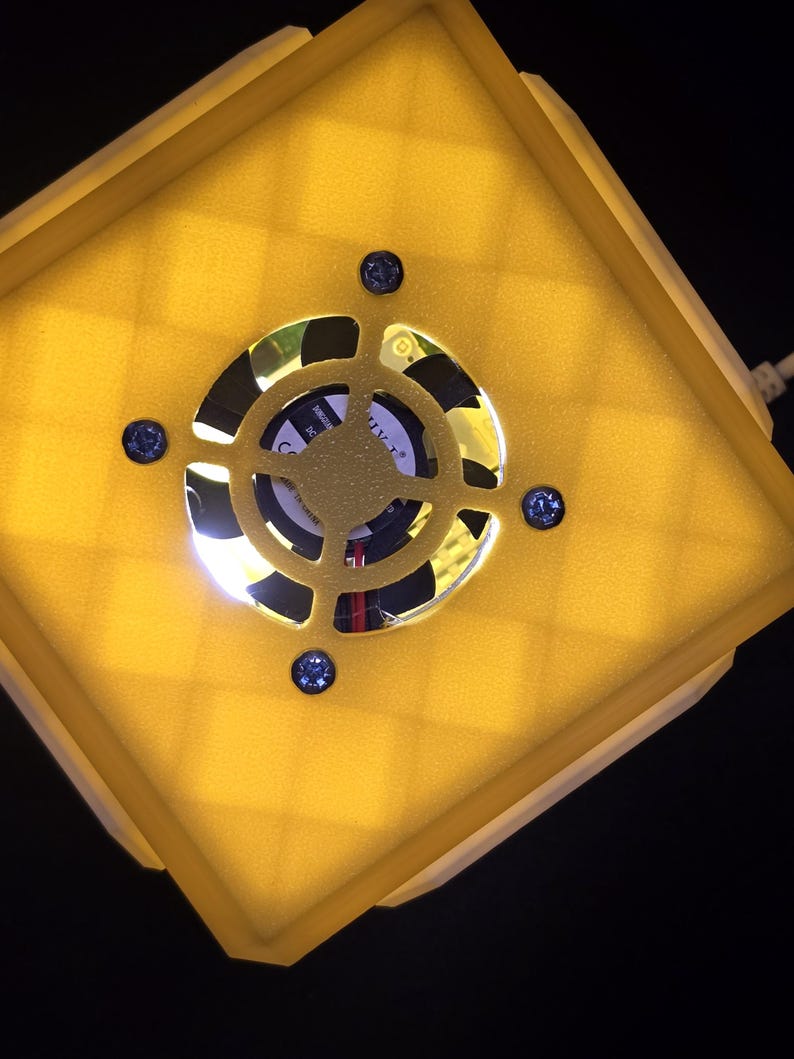 May include: A yellow square-shaped object with a white border and a black fan in the center. The fan has a white label with black text that reads "CUTL+ 24V 0.15A".