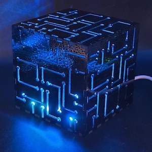 May include: A black, cube-shaped lamp with a circuit board design. The lamp emits a bright blue light through the cut-out patterns, creating a futuristic aesthetic. The lamp is plugged in with a white cable.