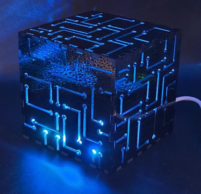 May include: A black cube-shaped lamp featuring intricate, laser-cut circuit board patterns on its exterior panels. The cutouts are illuminated from within by a bright blue light, creating a glowing, high-contrast effect across the surfaces.