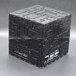 May include: A black cubic computer case featuring a laser-cut circuit board pattern on all visible sides. The enclosure has cutouts revealing internal ports, including USB and Ethernet connections, constructed from interlocking flat panels.