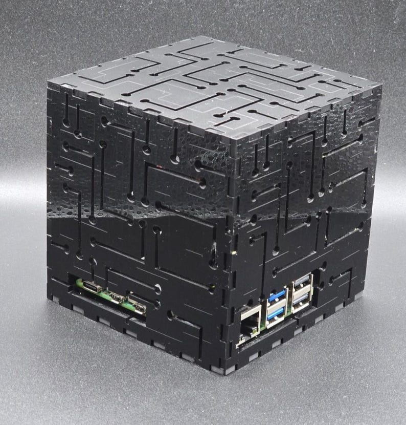 May include: A black cubic computer case featuring a laser-cut circuit board pattern on all visible sides. The enclosure has cutouts revealing internal ports, including USB and Ethernet connections, constructed from interlocking flat panels.