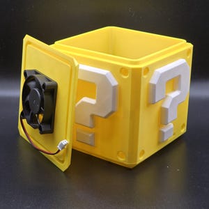 May include: A yellow plastic box with a question mark design on each side. The box is open and a small black fan is visible inside.