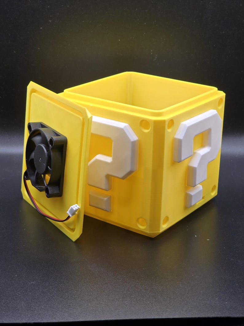 May include: A yellow plastic box with a question mark design on each side. The box is open and a small black fan is visible inside.
