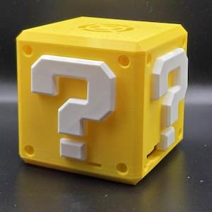 Könnte beinhalten: Ein gelber 3D-gedruckter Würfel mit einem weißen Fragezeichen auf jeder Seite. Der Würfel ist eine Nachbildung eines klassischen Videospielgegenstands.