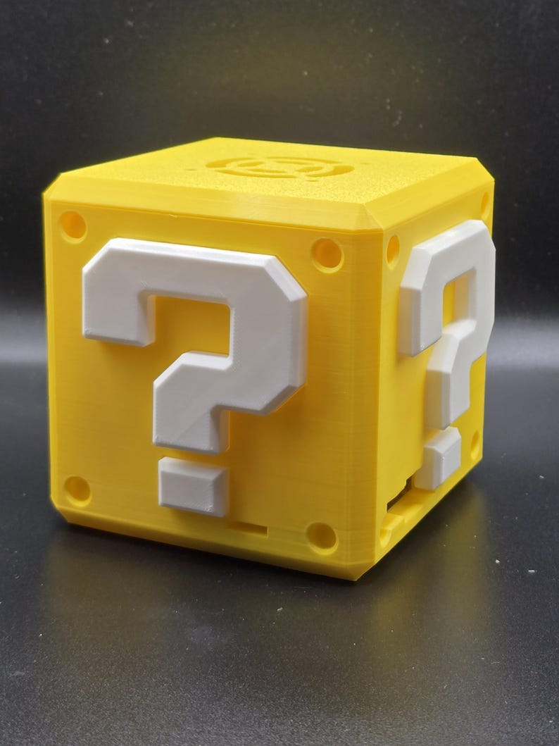 May include: A yellow 3D printed cube with a white question mark on each side. The cube is a replica of a classic video game item.
