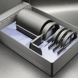 May include: An open, rectangular, gray case with a hinged lid, containing several black, curved objects and a cylindrical item. The interior has a blue and white patterned lining. The case is on a dark surface.