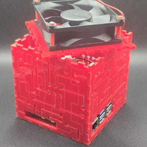 May include: A red, cube-shaped computer case featuring a laser-cut circuit board pattern on its exterior panels. A black cooling fan is mounted on the top panel, secured with orange spacers. The case reveals internal ports, including USB and Ethernet.