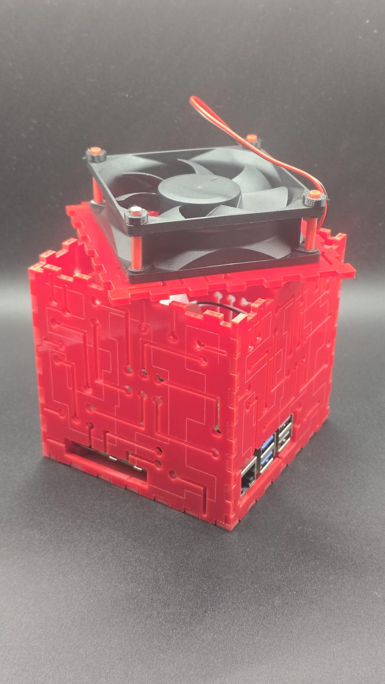 May include: A red, cube-shaped computer case featuring a laser-cut circuit board pattern on its exterior panels. A black cooling fan is mounted on the top panel, secured with orange spacers. The case reveals internal ports, including USB and Ethernet.