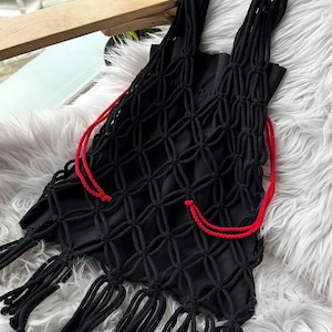 以下が含まれることがあります： 幾何学的なネットデザインと長いフリンジが付いた、黒いマクラメトートバッグ。赤いドローストリングと太い編み込みハンドルが特徴です。バッグは柔らかい織り素材で作られています。