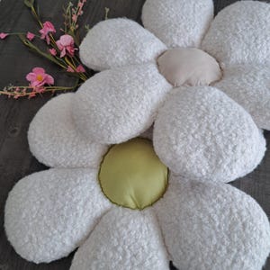 Könnte beinhalten: Ein großes, weißes, gänseblümchenförmiges Kissen mit einer strukturierten, flauschigen Oberfläche. Die Mitte ist hellbeige und die Mitte der Blume hellgrün. Rosa Blüten und grüne Stiele sind im Hintergrund.