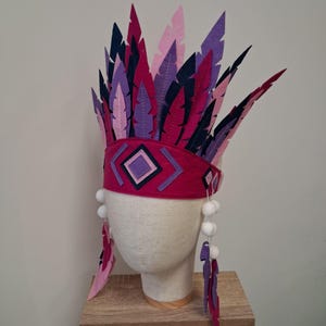 Peut inclure: Coiffe faite à la main avec un bandeau magenta orné de motifs géométriques et de pompons blancs. Elle est ornée de plumes en feutre roses, violettes et bleu marine, créant un affichage visuel saisissant.