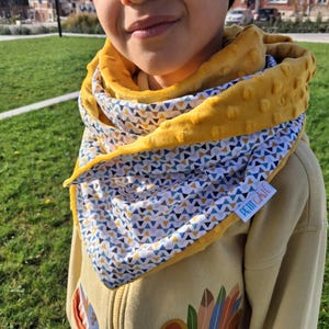 Peut inclure: Écharpe jaune et à motifs avec un motif géométrique. L'écharpe a un extérieur doux et texturé et une étiquette blanche avec le texte "PETITE GATE". L'écharpe est enroulée autour du cou.